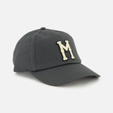 The Monogram Baseball Cap by MONCLER is a dark grey cap with a curved brim, featuring a large white "M" embroidered on the front.