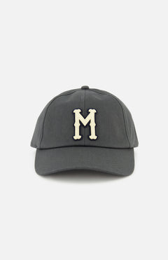 The MONCLER Monogram Baseball Cap features a grey design with a cream-colored embroidered letter M on the front, inspired by designer outerwear style, and is shown against a plain white background.
