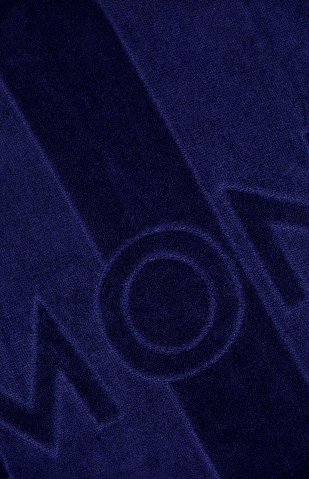 Close-up of the MONCLER Logo Beach Towel showing its textured dark blue surface, with bold raised and indented lettering—like "M" and "O"—and vertical stripe patterns.