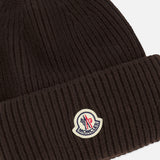The MONCLER Contrast Logo Beanie is a dark brown ribbed knit hat made from luxurious virgin wool, featuring a folded cuff and a sewn-on cream, red, and blue Moncler logo patch on the front.