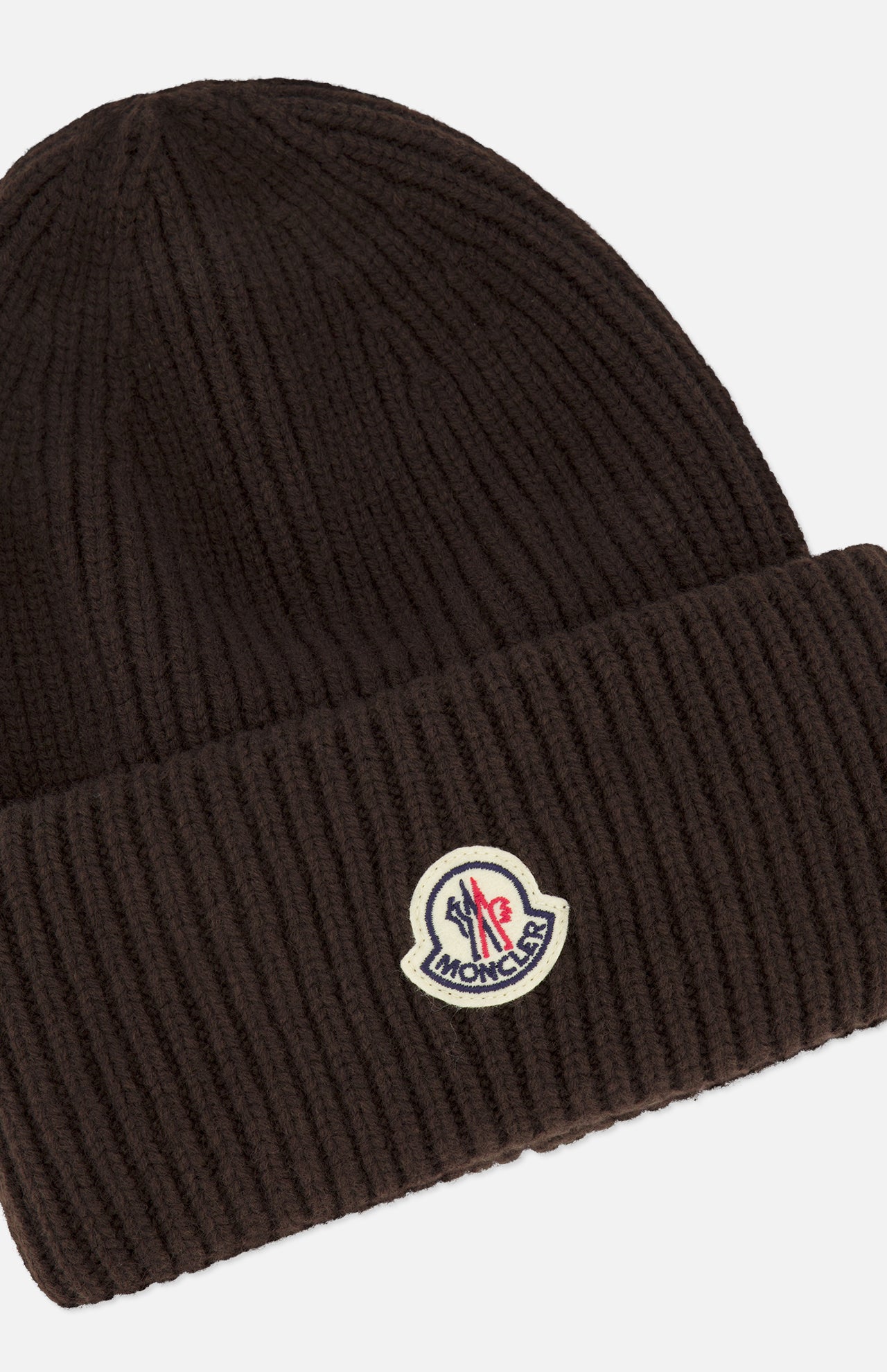 The MONCLER Contrast Logo Beanie is a dark brown ribbed knit hat made from luxurious virgin wool, featuring a folded cuff and a sewn-on cream, red, and blue Moncler logo patch on the front.