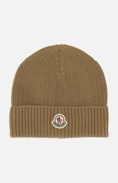 The MONCLER Ribbed Logo Beanie is a brown knit beanie with a folded cuff, featuring the signature Moncler patch on the front center. This stylish accessory is showcased against a plain white background.