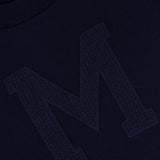 Close-up of a navy MONCLER Long Sleeve Crewneck T-Shirt featuring a large, textured "M" stitched on the front, with part of the label visible near the collar.