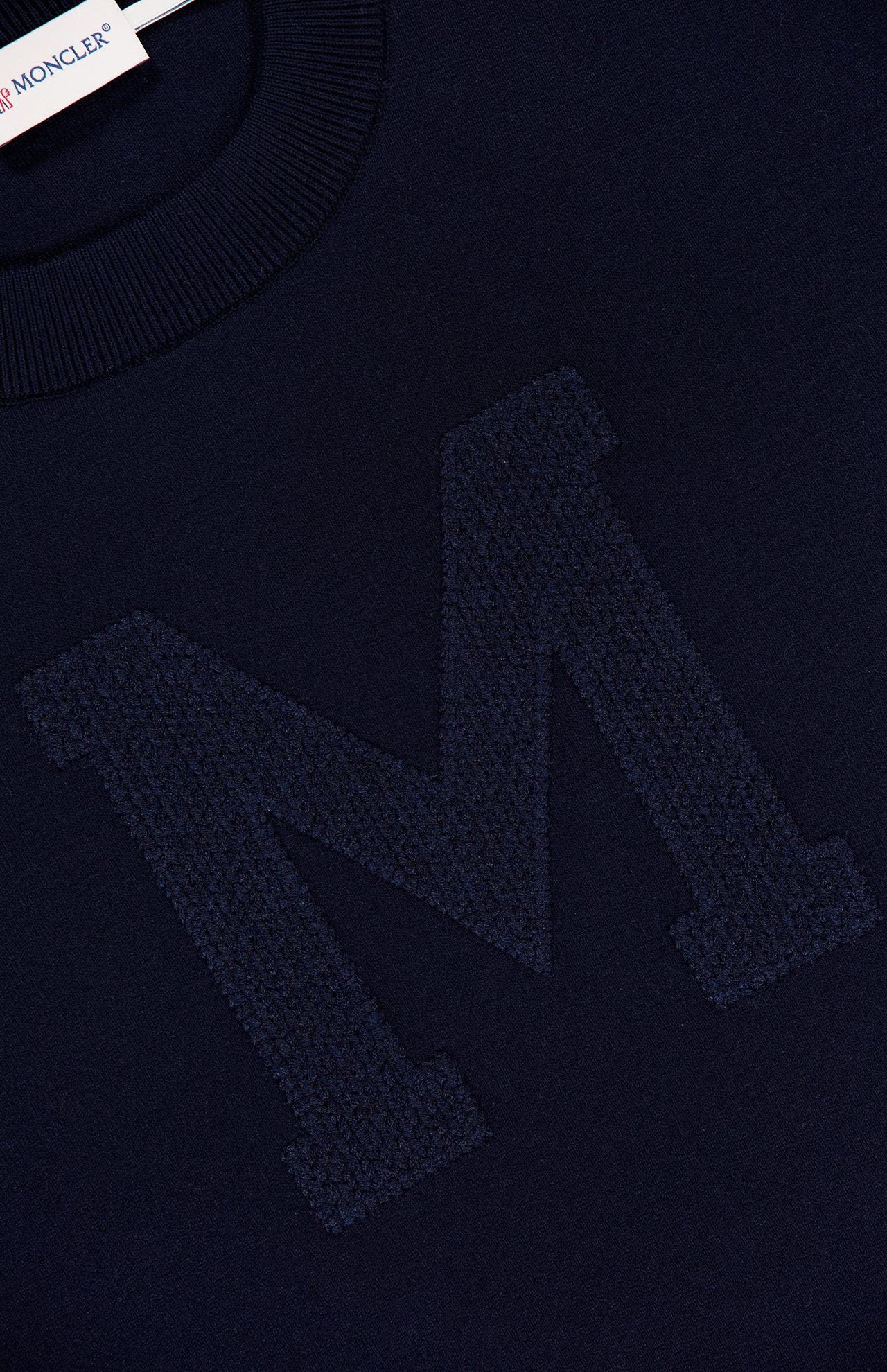 Close-up of a navy MONCLER Long Sleeve Crewneck T-Shirt featuring a large, textured "M" stitched on the front, with part of the label visible near the collar.