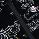 Close-up of the MONCLER Reversible Paisley Fleece Vest in black, showcasing a bold white and yellow paisley pattern and a prominently placed Moncler logo patch near the zippered seam—this eye-catching vest channels the playful charm of paisley teddy motifs while embodying the signature sophistication of MONCLER craftsmanship.