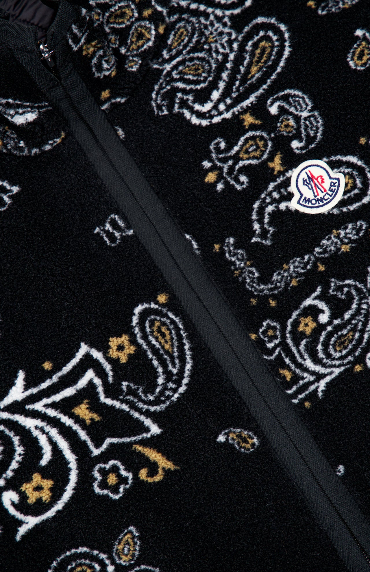 Close-up of the MONCLER Reversible Paisley Fleece Vest in black, showcasing a bold white and yellow paisley pattern and a prominently placed Moncler logo patch near the zippered seam—this eye-catching vest channels the playful charm of paisley teddy motifs while embodying the signature sophistication of MONCLER craftsmanship.
