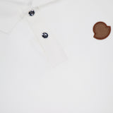 Close-up of the MONCLER Short Sleeve Cotton Polo Shirt in white cotton, featuring two dark buttons, a brown logo patch on the chest, and textured fabric with a partial view of the collar.