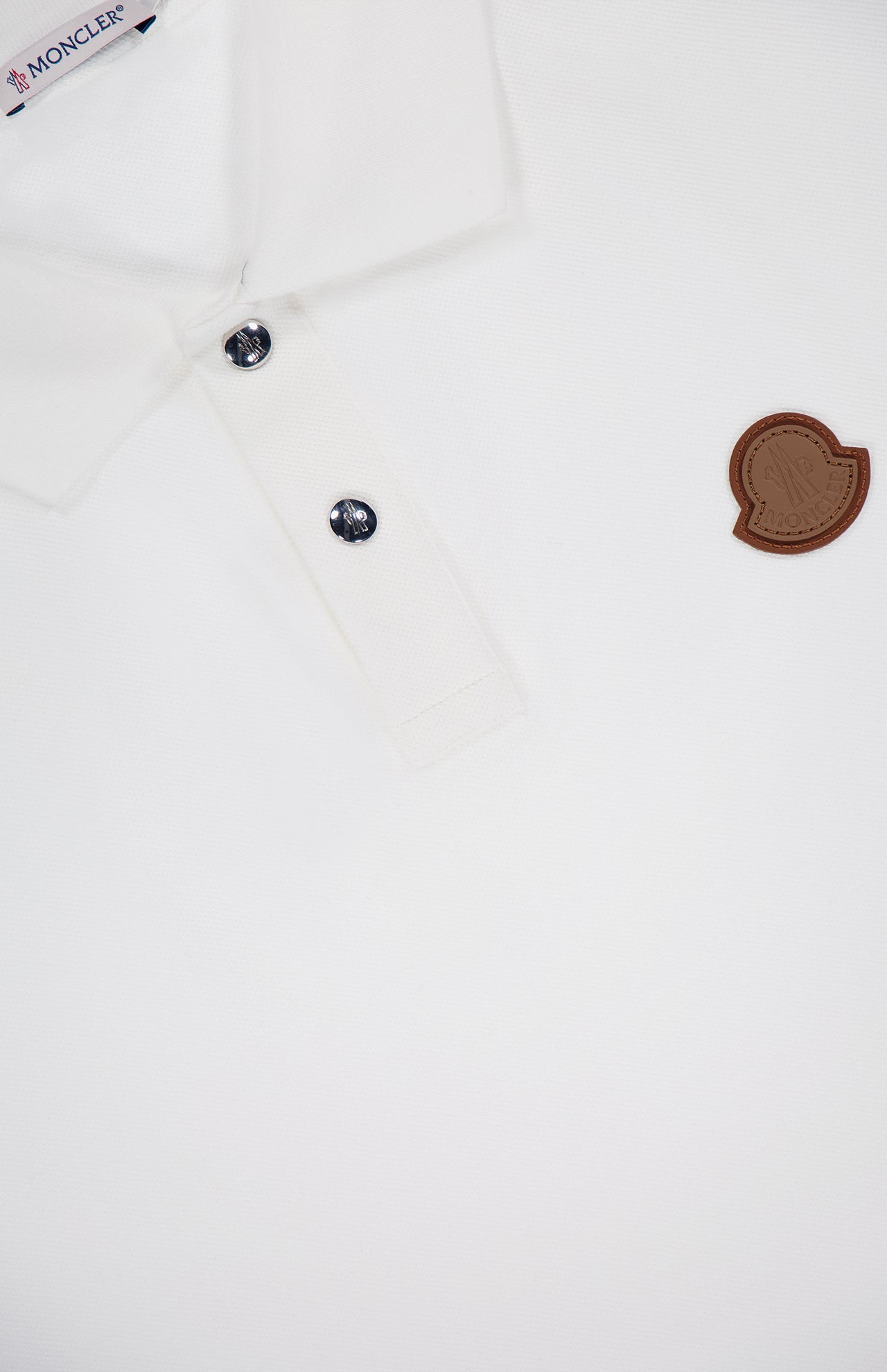 Close-up of the MONCLER Short Sleeve Cotton Polo Shirt in white cotton, featuring two dark buttons, a brown logo patch on the chest, and textured fabric with a partial view of the collar.