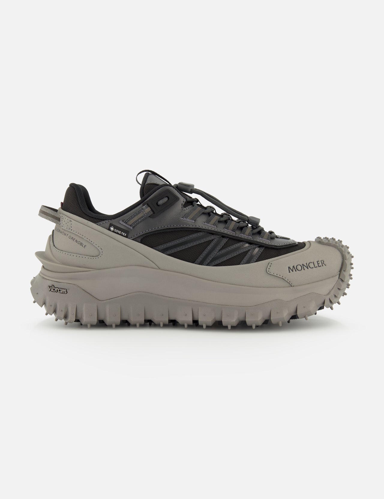 A side view of the MONCLER Trailgrip GTX Low Top Sneakers in gray and black, featuring a chunky Vibram tread sole for grip, a lace-up design, and the Moncler logo on the front side.