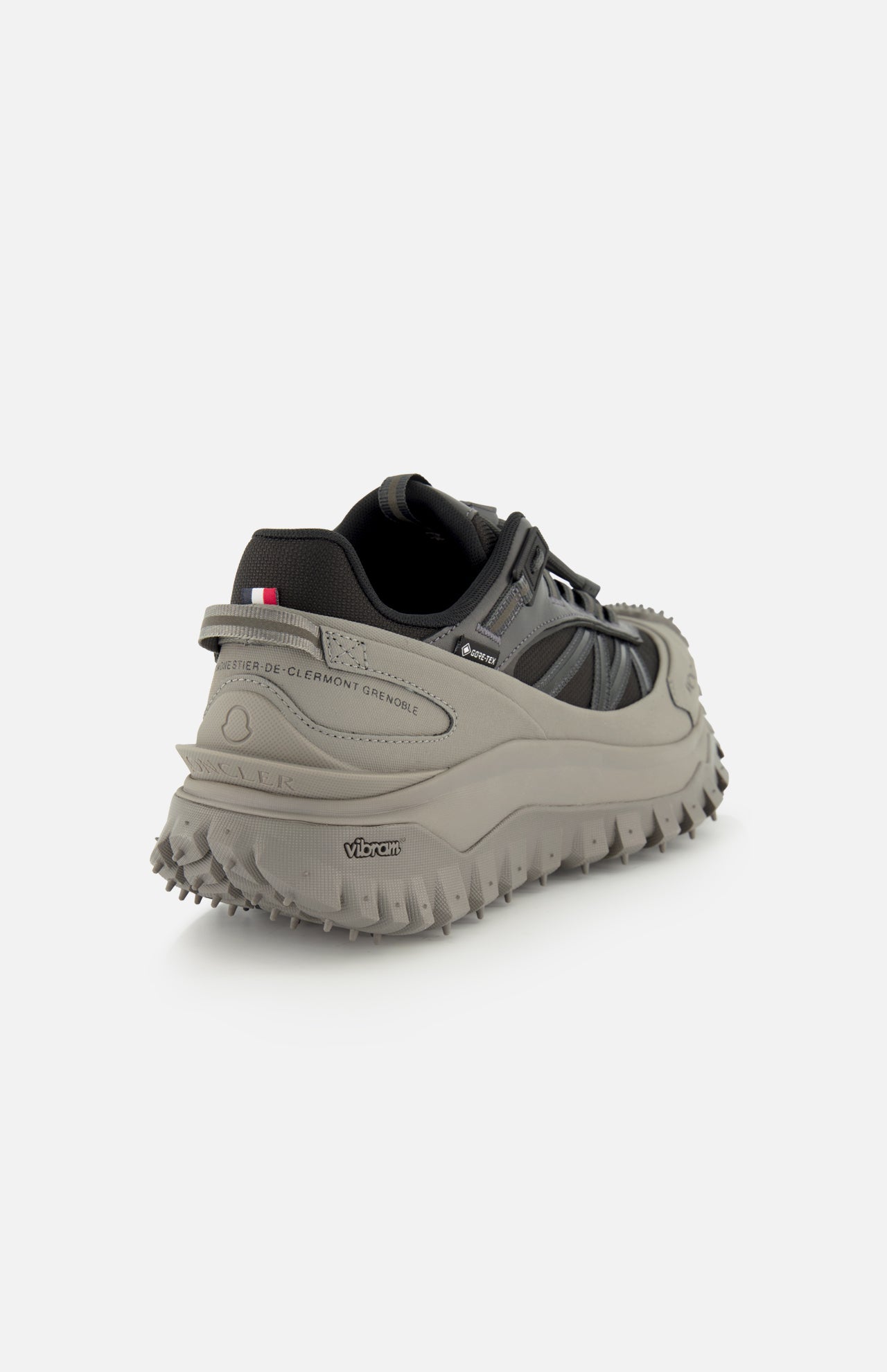 A close-up rear view of the MONCLER Trailgrip GTX Low Top Sneakers in gray and black, featuring a lugged Vibram sole, nylon upper, and a small French flag on the heel, set against a plain white background.