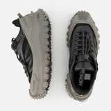 Two MONCLER Trailgrip GTX Low Top Sneakers in black and grey, with chunky Vibram soles—one viewed from the side, one from above—are displayed against a white background.