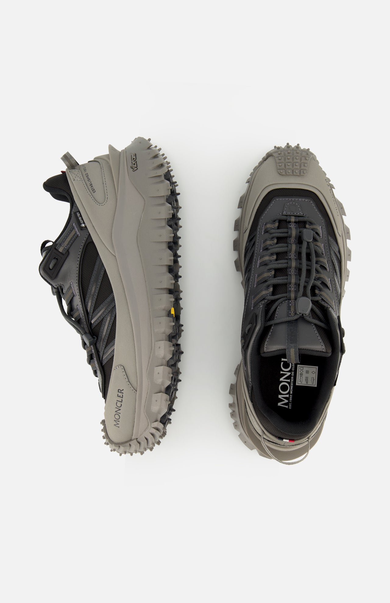 Two MONCLER Trailgrip GTX Low Top Sneakers in black and grey, with chunky Vibram soles—one viewed from the side, one from above—are displayed against a white background.