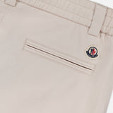 Close-up of MONCLER Drawstring Stretch Cotton Trousers highlights a horizontal back pocket, belt loops, elastic waistband with drawstring, and a small circular Moncler logo patch above the pocket.