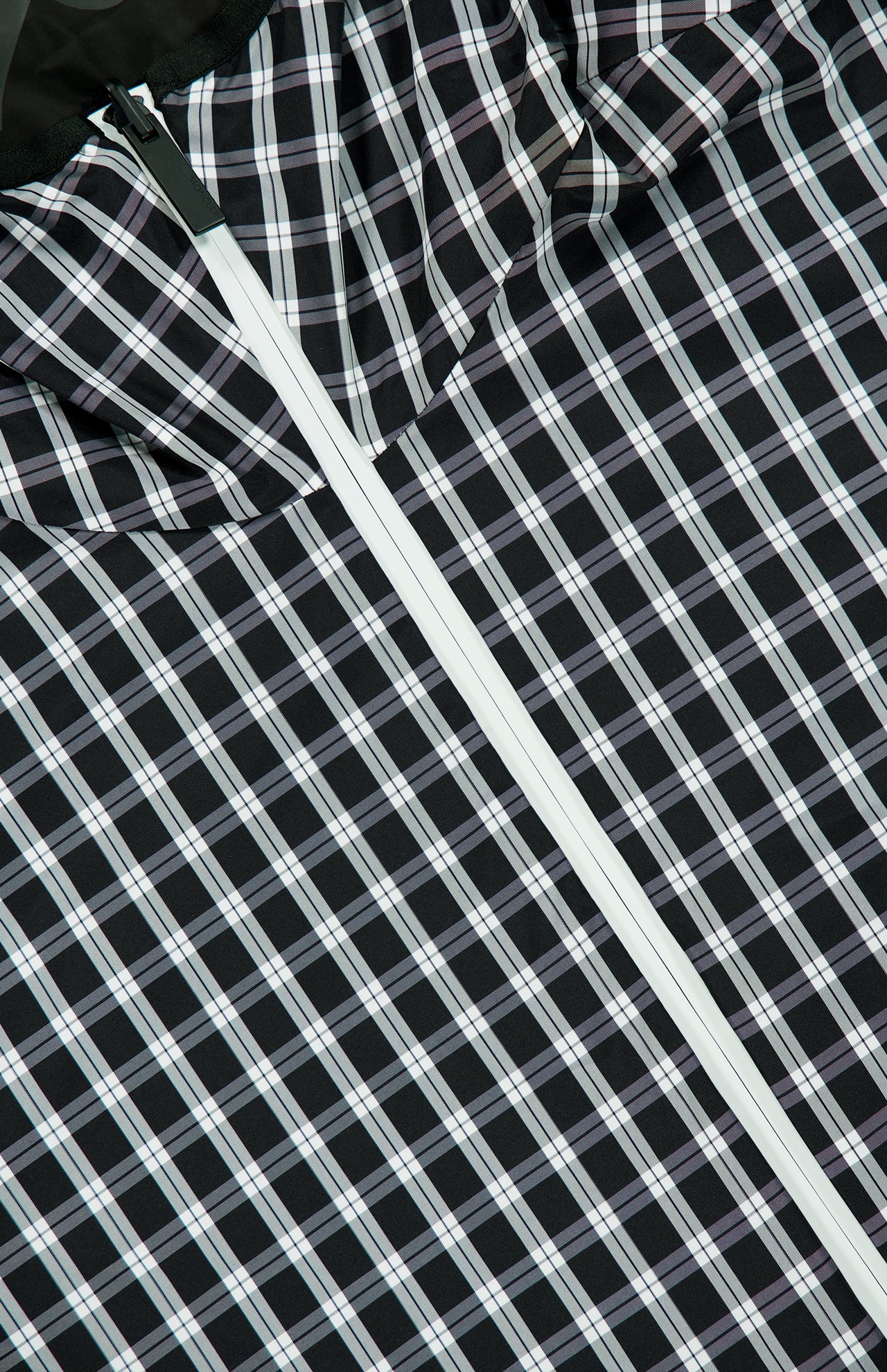 A close-up of the black fabric with a white and light blue plaid pattern, partially folded, reveals a white diagonal zipper—indicating this is likely part of the MONCLER Vandelli Reversible Plaid Jacket.