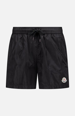 The MONCLER Drawstring Swim Trunk is a men's black technical nylon short with an elastic waistband, drawstring closure, side pockets, and a small embroidered logo patch on the left leg, shown against a plain white background.