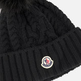 A black MONCLER Cableknit Pom Beanie featuring a large, fluffy faux fur pom pom on top and a MONCLER logo patch on the folded brim.