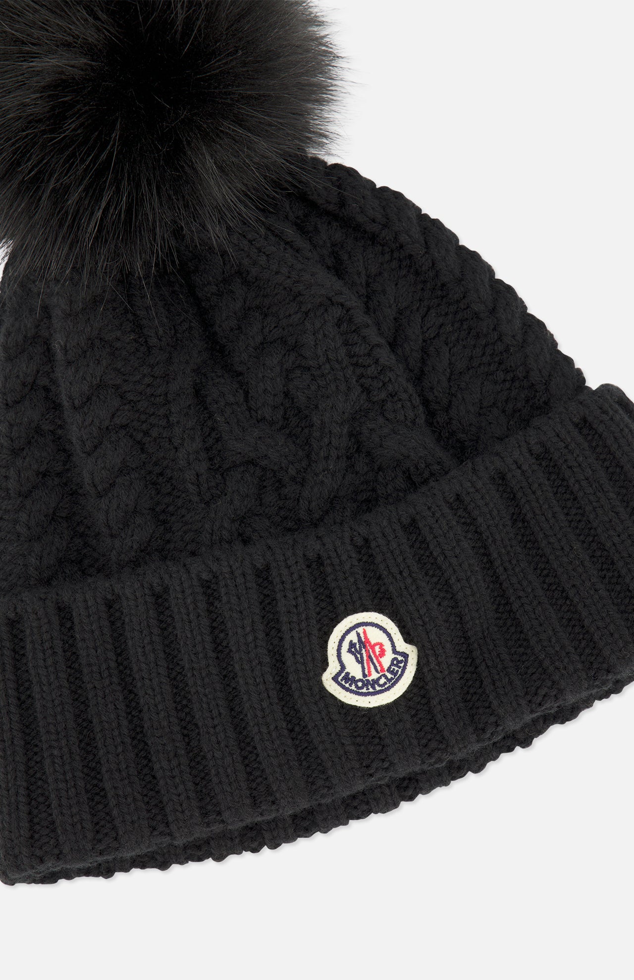 A black MONCLER Cableknit Pom Beanie featuring a large, fluffy faux fur pom pom on top and a MONCLER logo patch on the folded brim.
