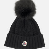 A black MONCLER Cableknit Pom Beanie featuring a large faux fur pom pom on top, a ribbed folded brim, and a round Moncler logo patch.