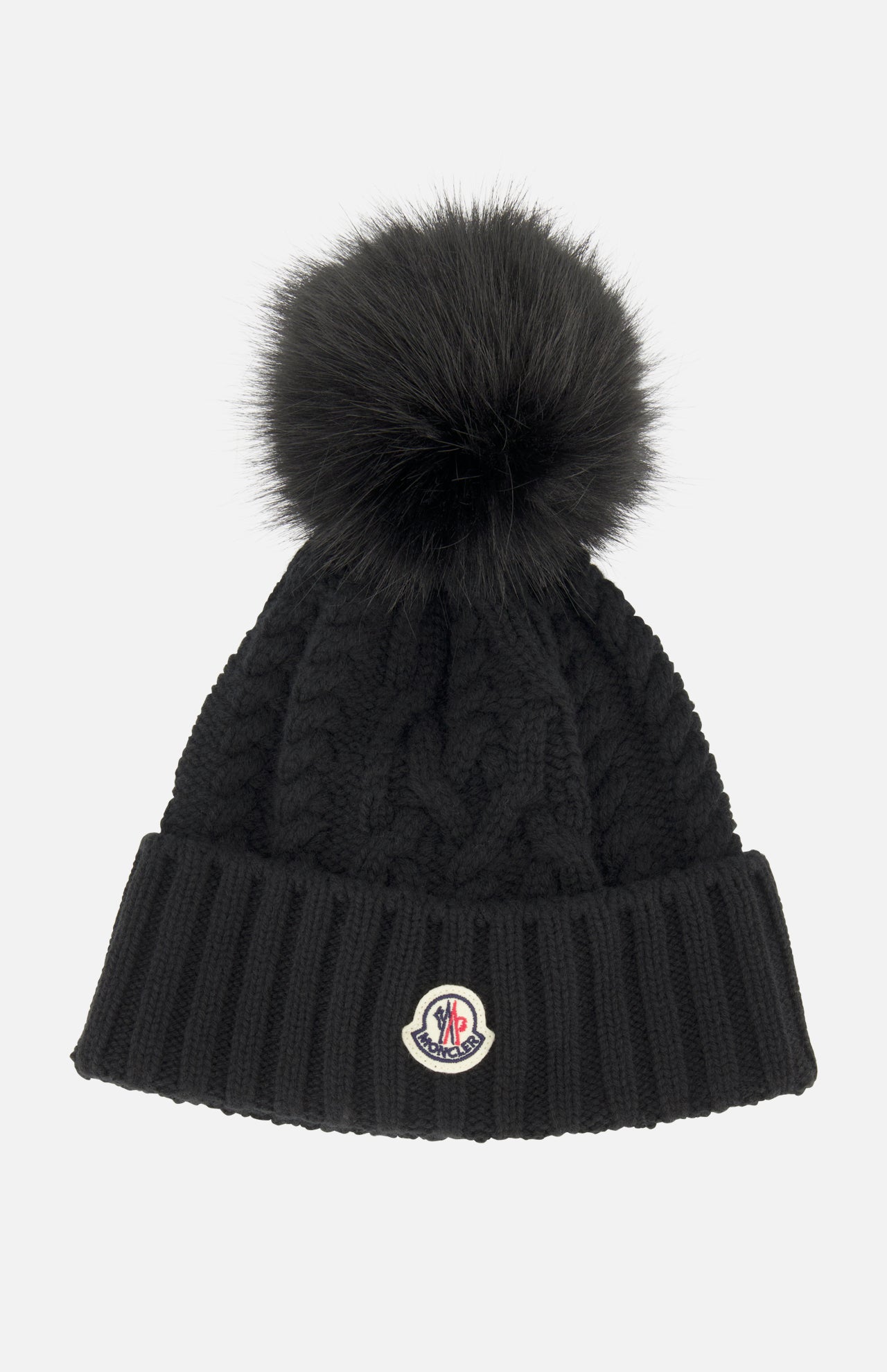 A black MONCLER Cableknit Pom Beanie featuring a large faux fur pom pom on top, a ribbed folded brim, and a round Moncler logo patch.