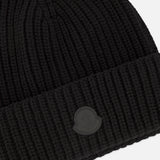 The MONCLER Tonal Logo Beanie is crafted from premium virgin wool cashmere in black, featuring a ribbed knit design with a folded cuff and a subtle round logo patch on the front. Showcasing a chunky texture, this beanie offers a simple and classic aesthetic.