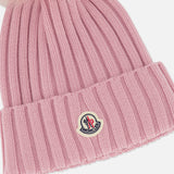The MONCLER Ribbed Pom Beanie is a pink ribbed knit hat featuring a large fluffy faux fur pom pom on top and a Moncler logo patch sewn onto the folded brim.