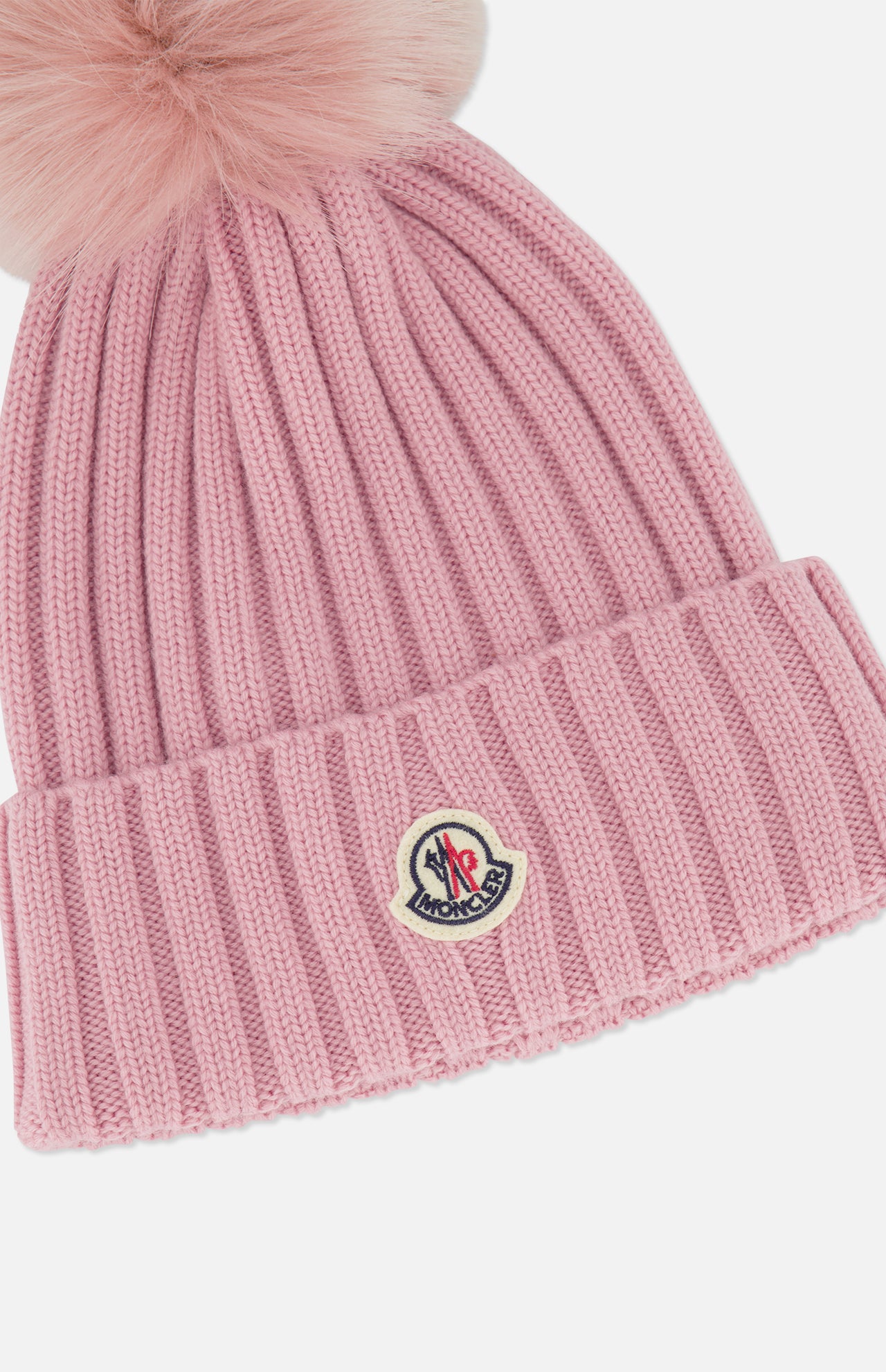 The MONCLER Ribbed Pom Beanie is a pink ribbed knit hat featuring a large fluffy faux fur pom pom on top and a Moncler logo patch sewn onto the folded brim.