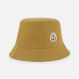 A mustard yellow MONCLER Reversible Bucket Hat with visible stitching and a small embroidered logo patch on the front, shown on a plain white background—blending style and MONCLER's renowned outerwear heritage.