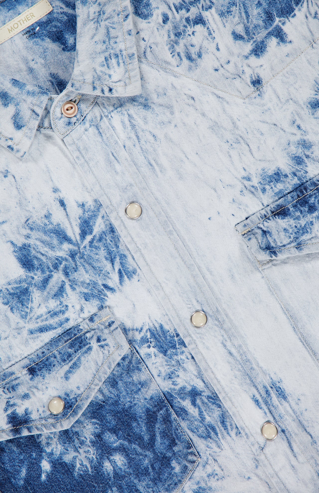 Close-up of The Exes And Ohs Overshirt by MOTHER, featuring a blue and white bleached tie-dye pattern, silver snap buttons, chest pockets, and a fabric label in the top left—showcasing its Western-inspired style.