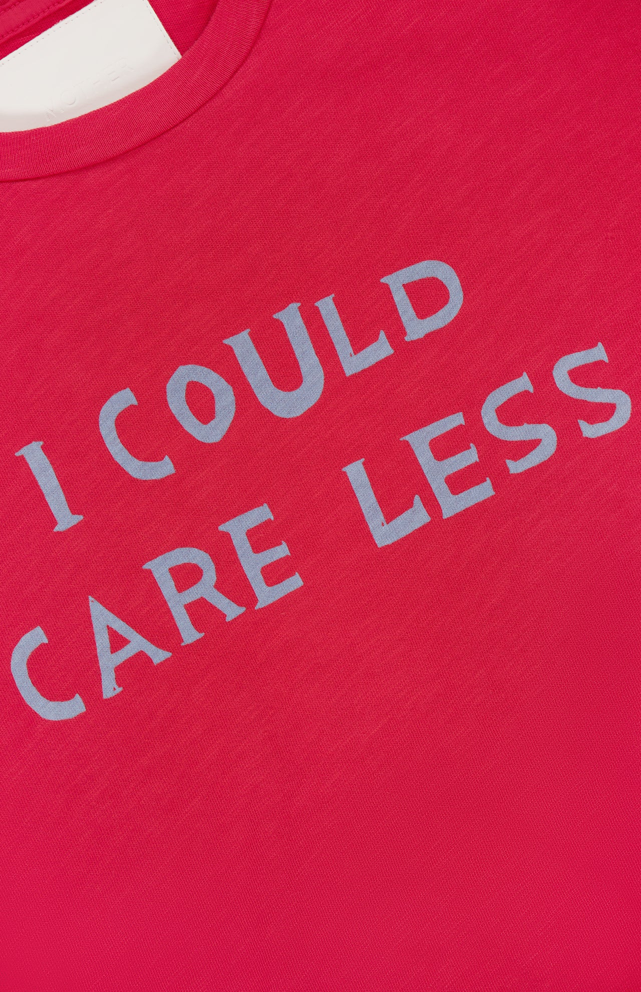 A close-up of The Sinful Slim T-shirt by MOTHER, a vintage-look red tee made from 100% cotton, featuring "I COULD CARE LESS" printed in bold light gray letters.