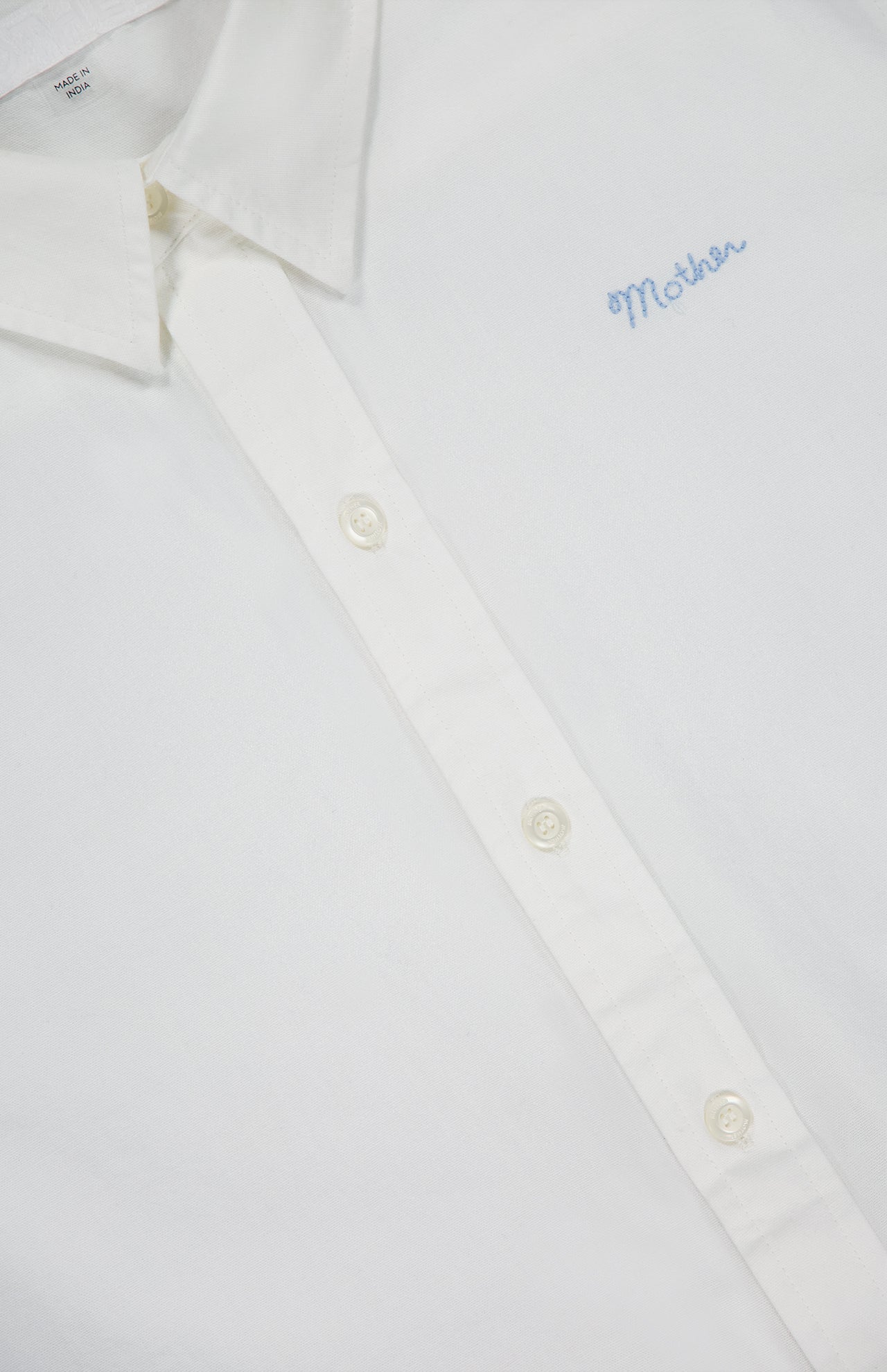 Close-up of The Bestie Button-front Shirt by MOTHER, a 100% cotton white shirt featuring cream-colored buttons and "Mother" embroidered in light blue on the chest.