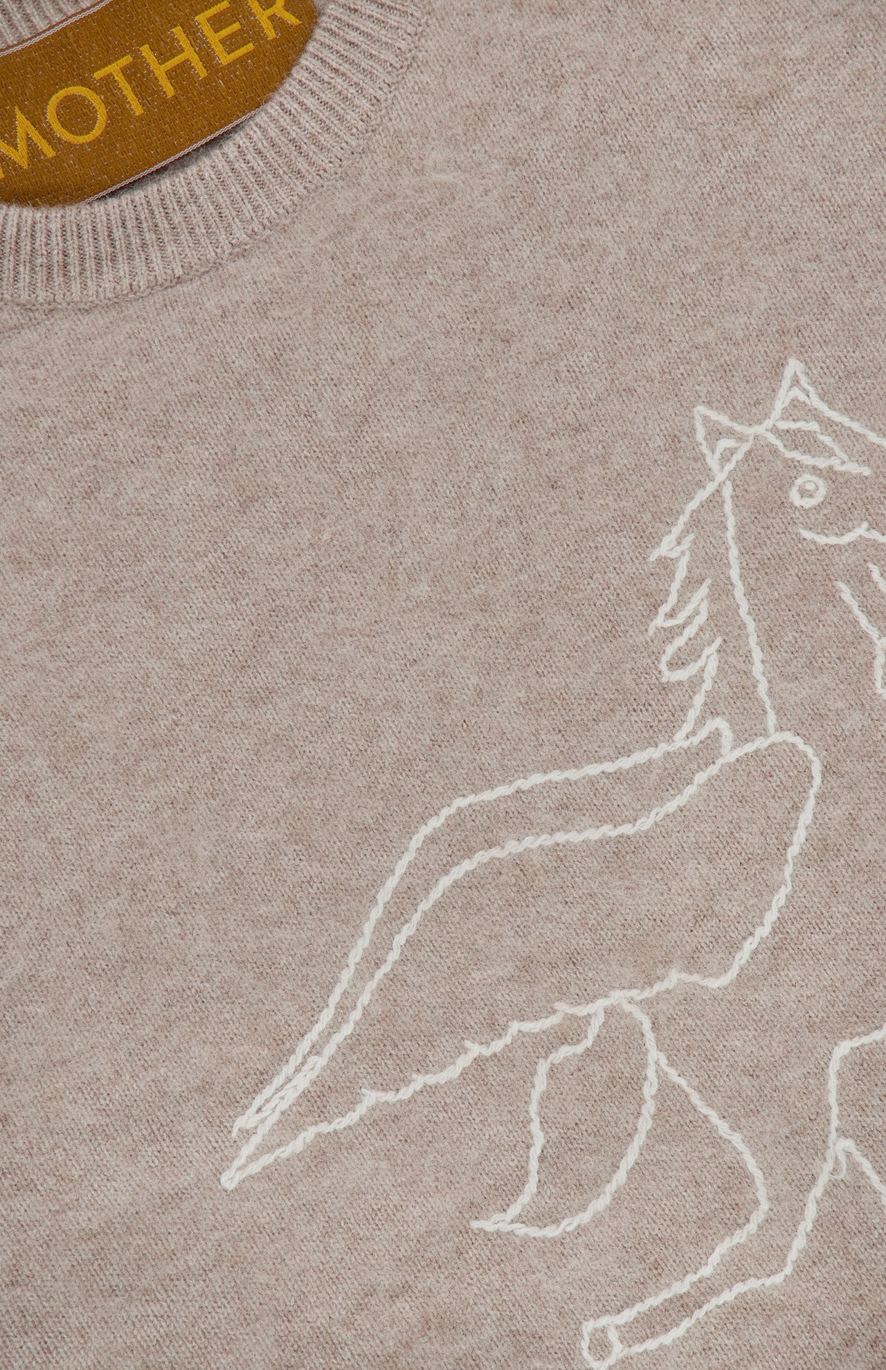A close-up of The Biggie Pegasus Jumper Sweater by MOTHER showcases its beige knit fabric, adorned with a partial white outline drawing of a winged horse. A brown collar tag displays the brand name, MOTHER, in yellow letters.