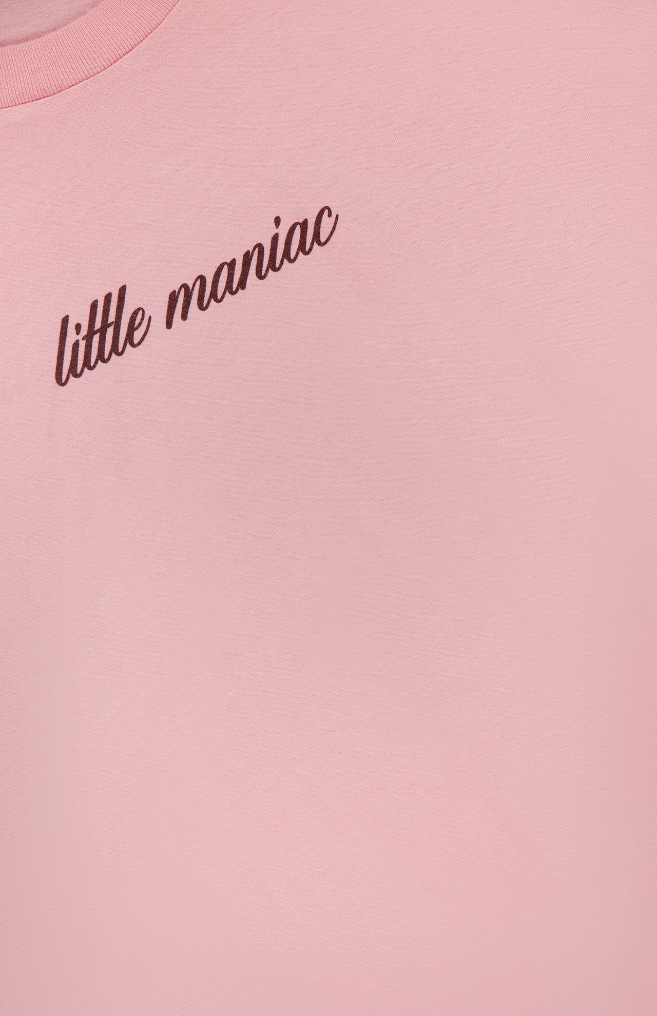 A close-up of MOTHER's The Boxy Goodie Goodie T-shirt in light pink, featuring "little maniac" in cursive brown text on the upper left side.