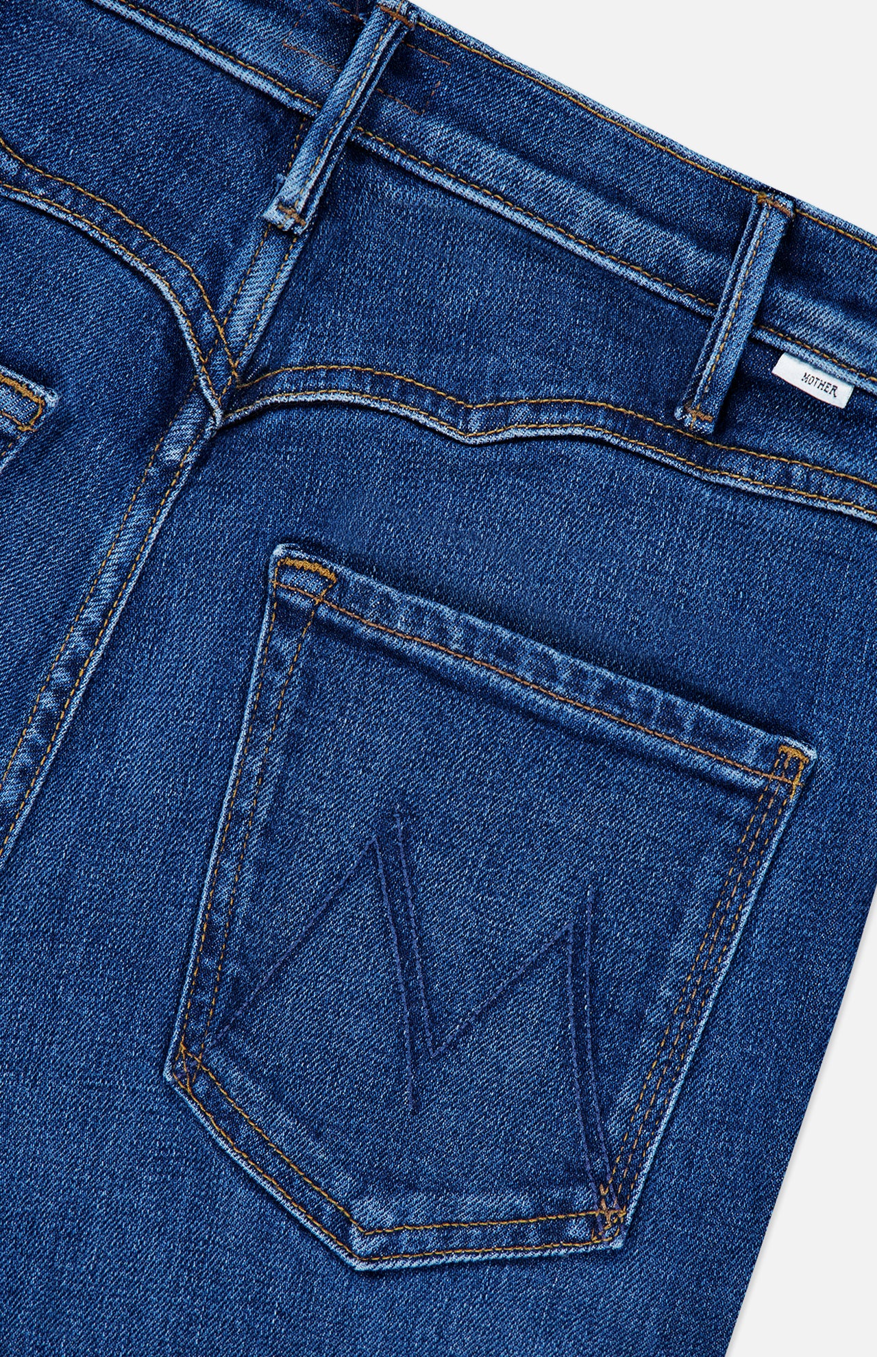 Close-up of the back of The Buckle Bunny Hustler Jean in Cosmic Cowgirl by MOTHER, featuring blue stretch denim with a stitched M design on the pocket, visible belt loops, and a small white label near the waistband.