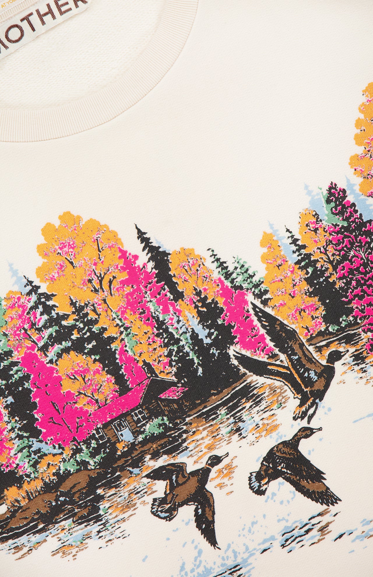 The Camper Lake Scene Sweatshirt by MOTHER features a lakeside cabin with autumn trees in pink, yellow, and orange. Three ducks fly over the water on this colorful cotton crew neck, with "MOTHER" partly visible at the neckline.