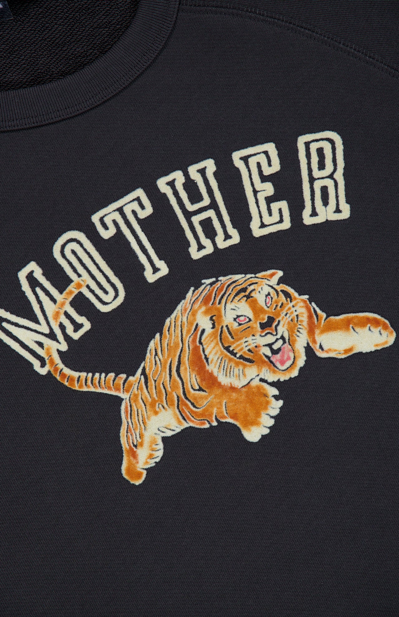 A dark 100% cotton sweatshirt called the Concert Tiger Sweatshirt by MOTHER, featuring the brand name in large letters above an illustrated orange and black tiger mid-leap with its mouth open and front paw extended.