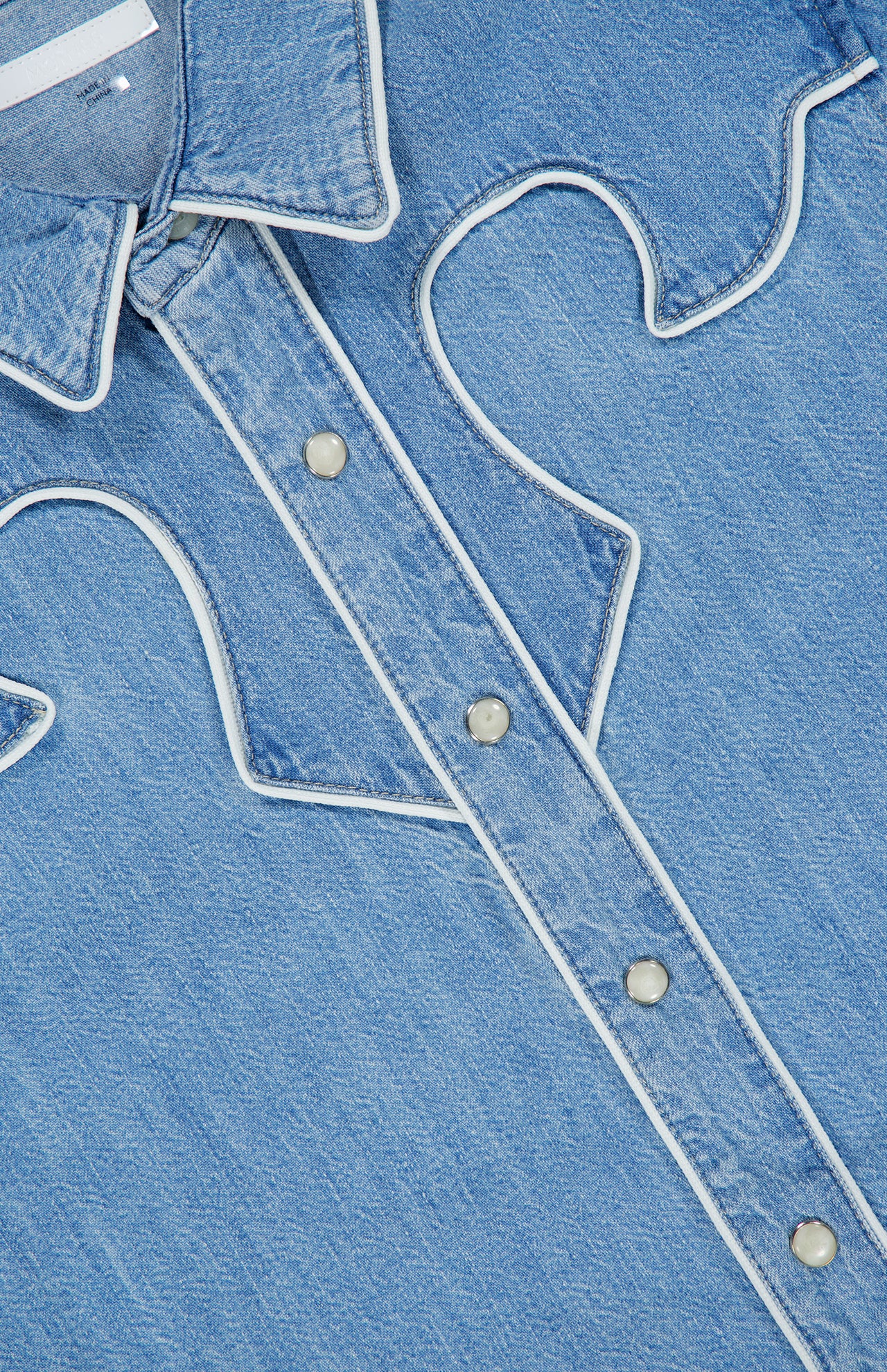 Close-up of The Country Exe’s Shirt by MOTHER, an oversized denim shirt featuring white piping details, silver snap buttons, and a pointed collar. Decorative stitching forms a western-inspired pattern on the front, channeling the timeless charm of a classic cowboy shirt.