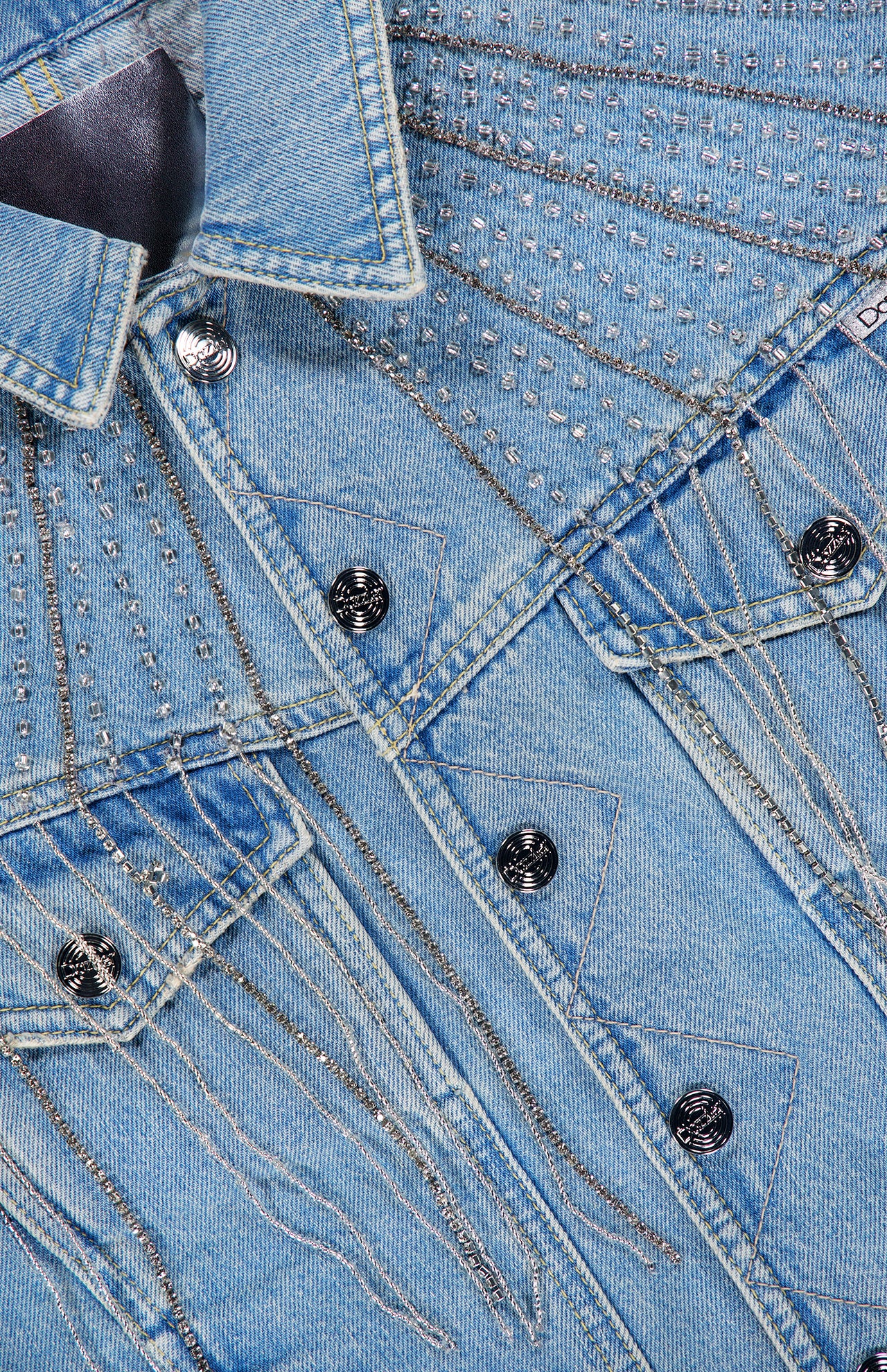 Close-up of the MOTHER Rhinestone Fringe Fly Away Jacket in light blue denim, adorned with decorative silver studs, metallic chain embellishments, and dazzling rhinestone fringe. Featuring metal buttons and detailed stitching, this jacket is the perfect statement piece for any party.