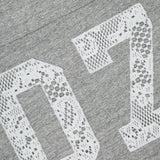 Close-up of The Footballer Lace T-shirt by MOTHER, a gray football jersey with a large white lace-style "07" print and a partially visible neckline tag featuring the brand’s cartoon face and "MOTHER.