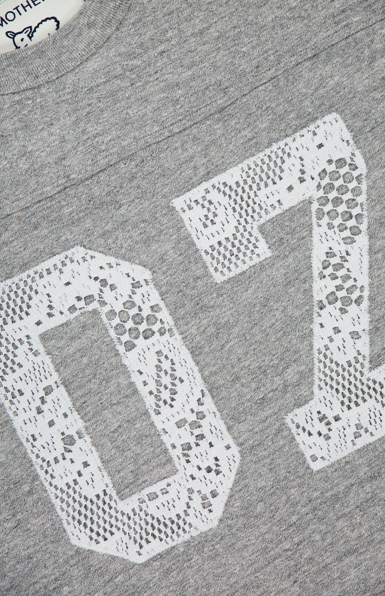 Close-up of The Footballer Lace T-shirt by MOTHER, a gray football jersey with a large white lace-style "07" print and a partially visible neckline tag featuring the brand’s cartoon face and "MOTHER.