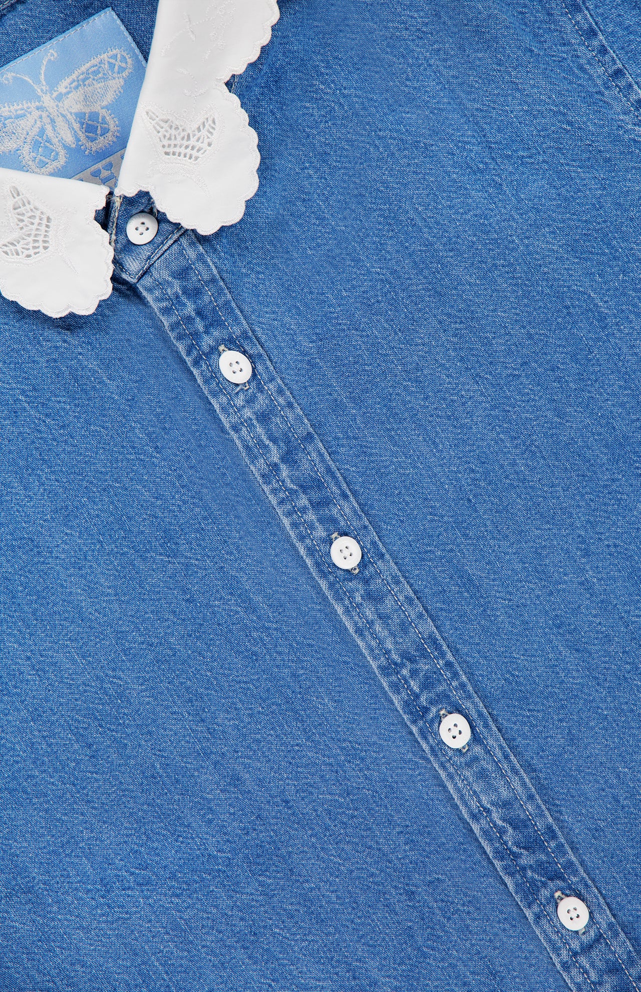 Close-up of The Frilly Foxy Boxy Denim Shirt by MOTHER: a blue 100% cotton button-down with white buttons and a scalloped, butterfly-embroidered collar featuring delicate white trim.