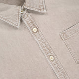 A close-up of The Honor Roll Cropped Shirt by MOTHER—a women’s button-up beige shirt with a pointed collar, chest pocket, four cream buttons, visible stitching, and a chic cropped hem.