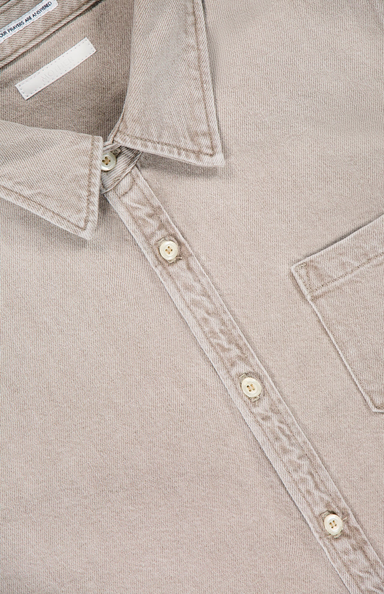 A close-up of The Honor Roll Cropped Shirt by MOTHER—a women’s button-up beige shirt with a pointed collar, chest pocket, four cream buttons, visible stitching, and a chic cropped hem.