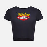Introducing The Itty Bitty Ringer T-shirt by MOTHER—a stylish black shrunken crew neck tee with short sleeves and a cropped hem. This eco-friendly top is crafted from recycled cotton and features a bold graphic of red lips with white teeth, topped with the word "MOTHER" in yellow text above the lips.