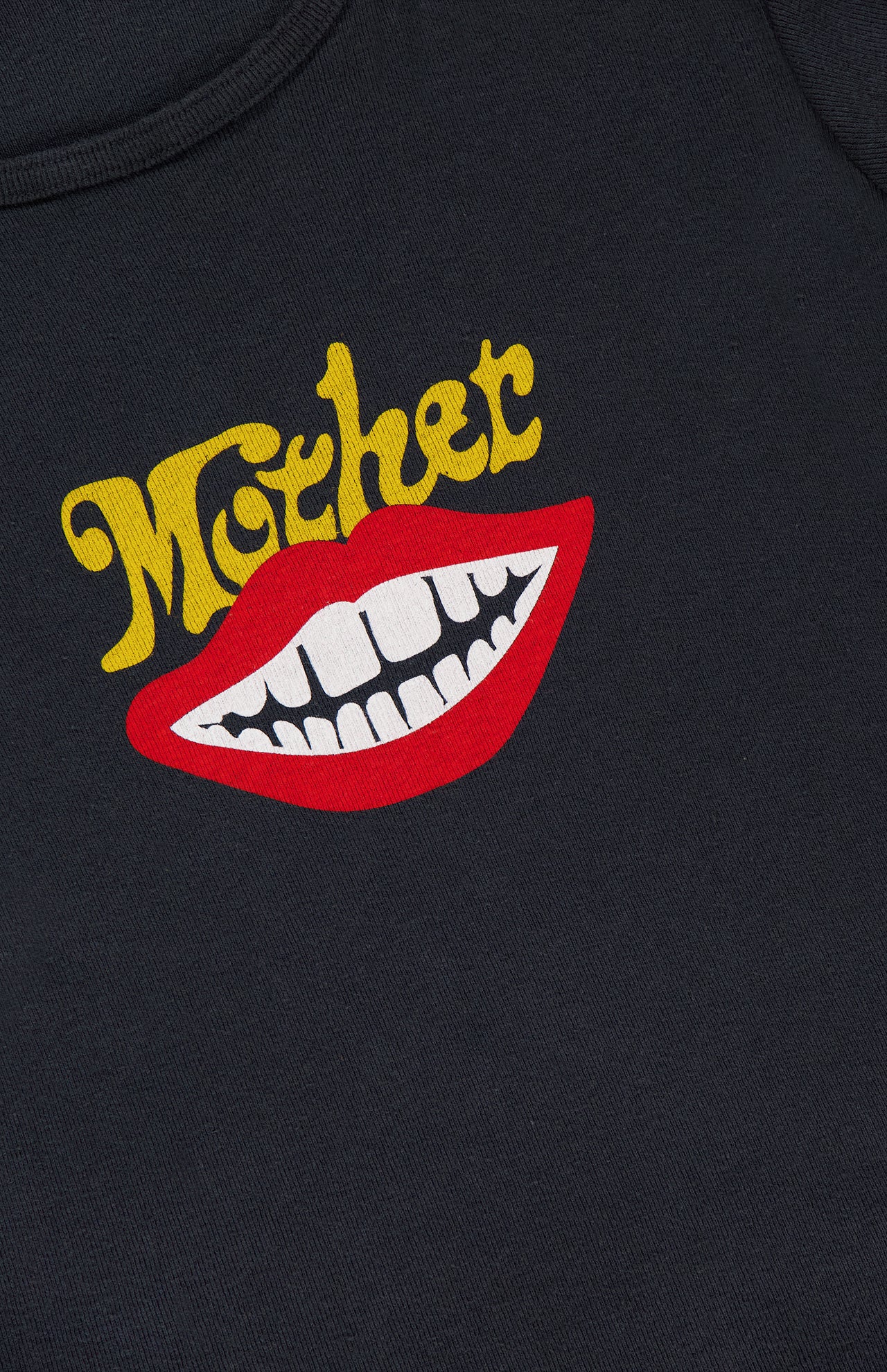 A graphic of red lips with white teeth is printed below the word MOTHER in yellow, stylized text on a dark fabric background. The Itty Bitty Ringer T-shirt by MOTHER features a shrunken crew neck, cropped hem, and is crafted from recycled cotton, blending bold design with sustainable style.