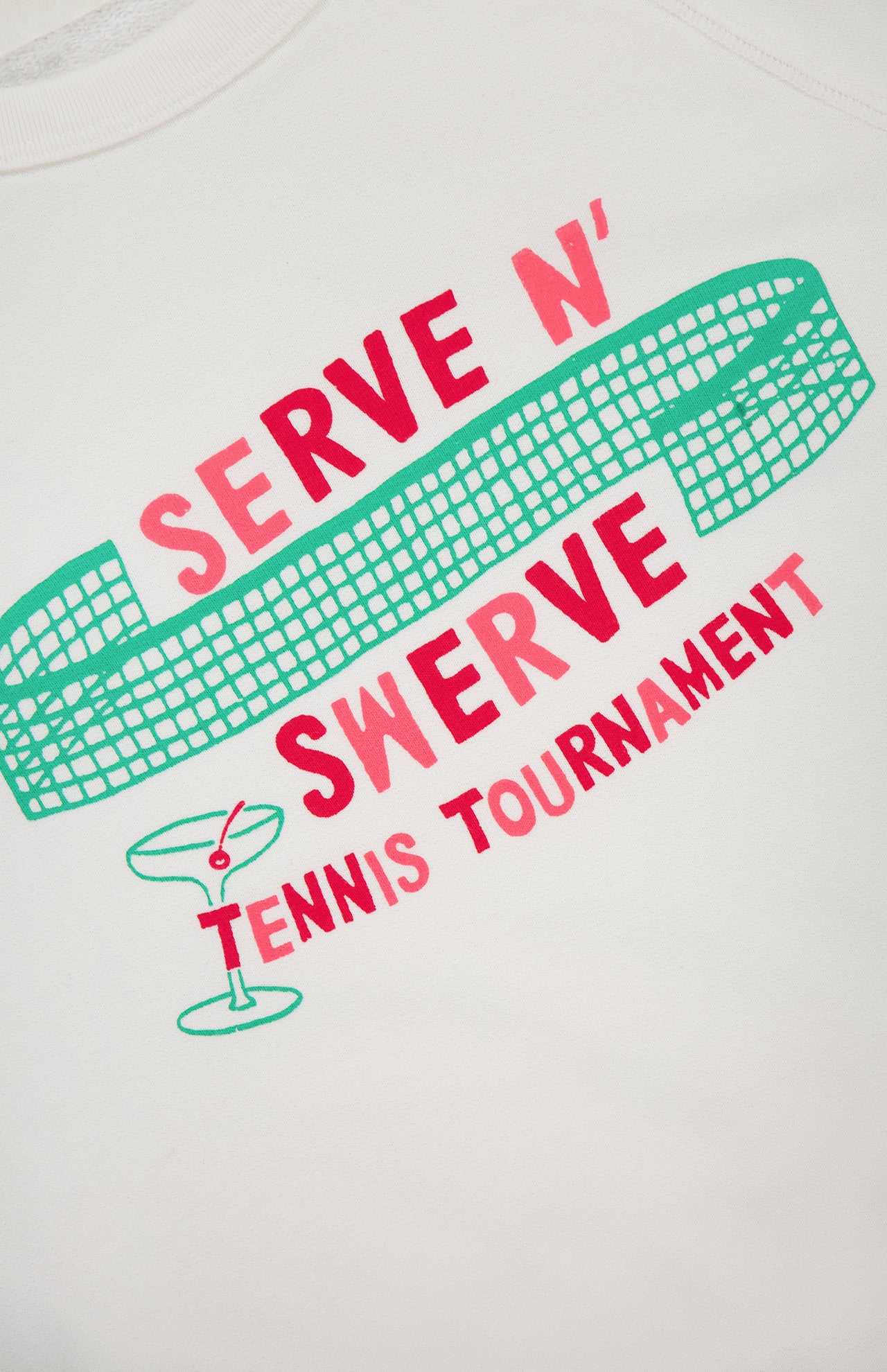 White background with a green tennis net illustration, red and pink "Serve N Swerve Tennis Tournament" text, and a green martini glass. The Itty Concert Tennis Sweater by MOTHER is a 100% cotton cropped raglan sweatshirt.