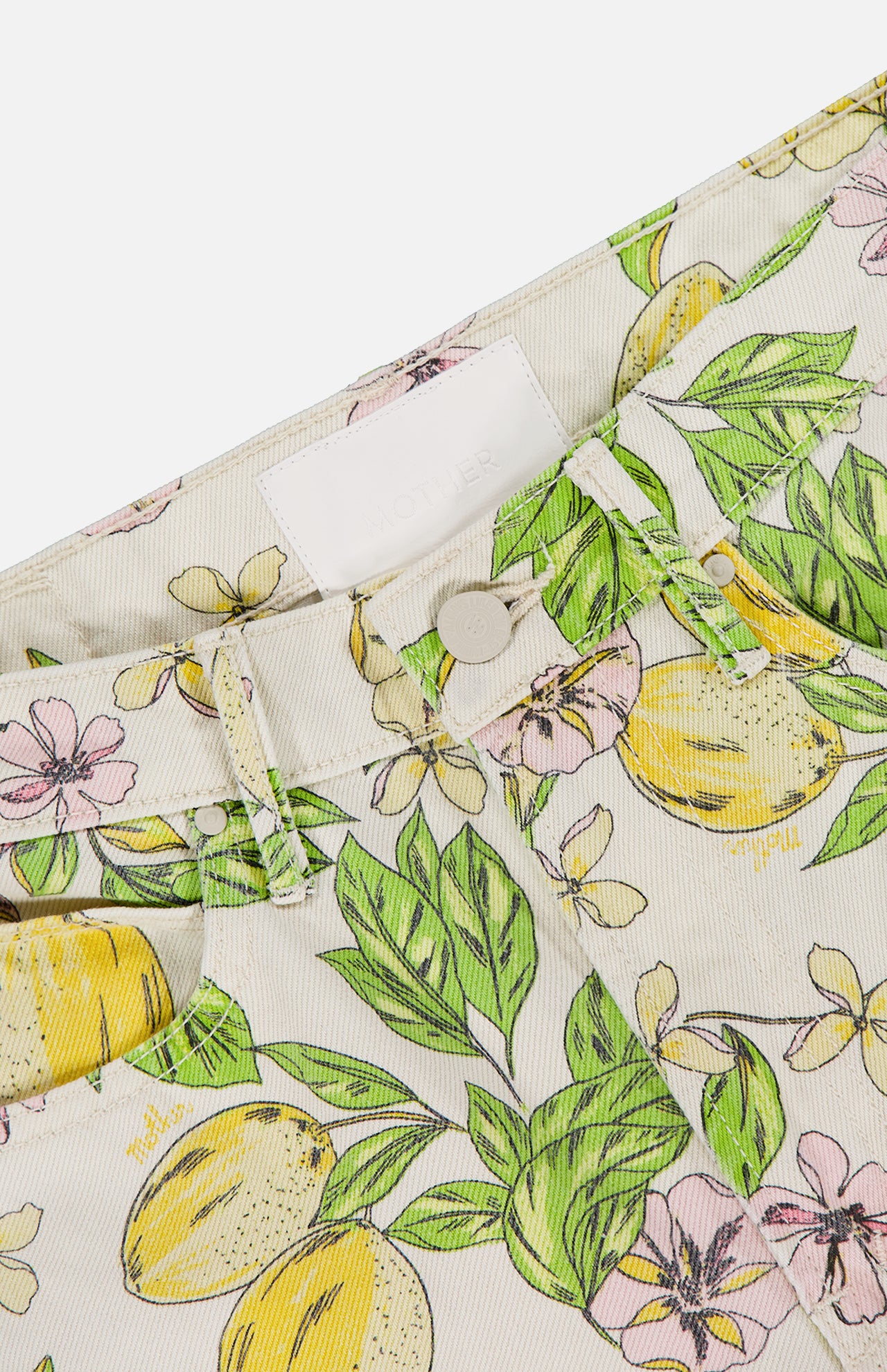 Close-up of The Kick It Pant in Lemonade Stand by MOTHER, featuring pastel lemon blossom print on rigid cotton denim, high-rise waistband, button detail, partial front pocket, and an empty white label on a light background.