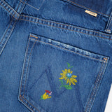 Close-up of MOTHER's The Rerun Zip Sneak Floral Print Jeans featuring high-waisted blue denim with painterly yellow and red floral embroidery on the pocket, a beige waistband tag, and visible stitching throughout.