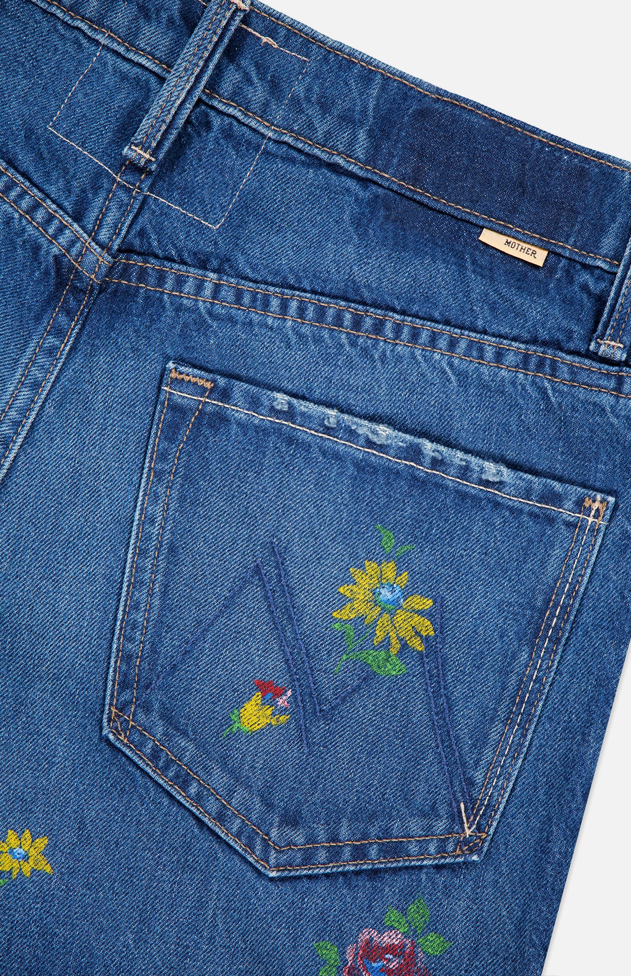 Close-up of MOTHER's The Rerun Zip Sneak Floral Print Jeans featuring high-waisted blue denim with painterly yellow and red floral embroidery on the pocket, a beige waistband tag, and visible stitching throughout.