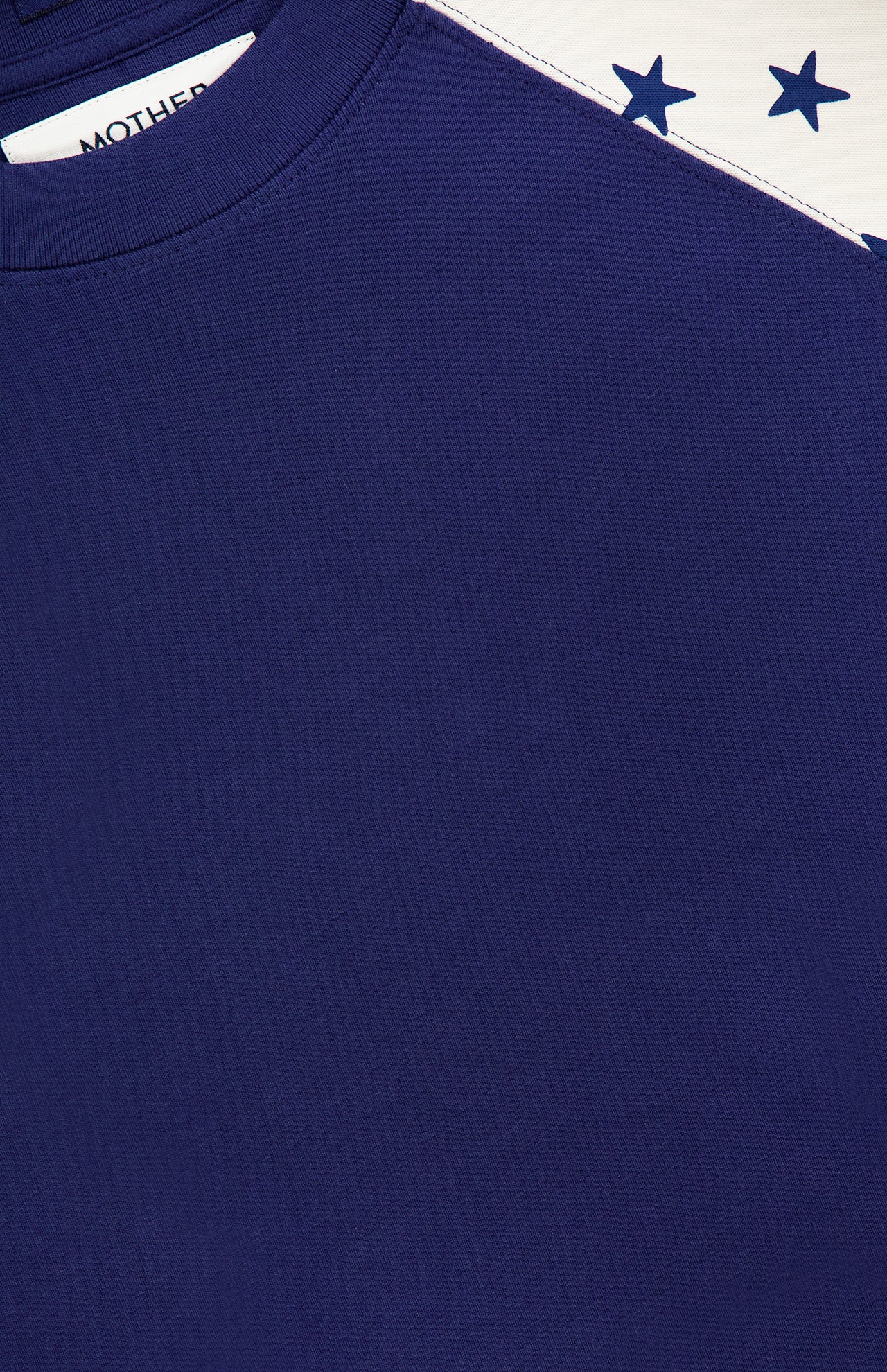 Close-up of The Concert Race T-shirt by MOTHER: a navy blue raglan tee with a white shoulder panel covered in dark blue stars. Made from 100% cotton for comfort and Americana style, with partial MOTHER label visible at the top left.