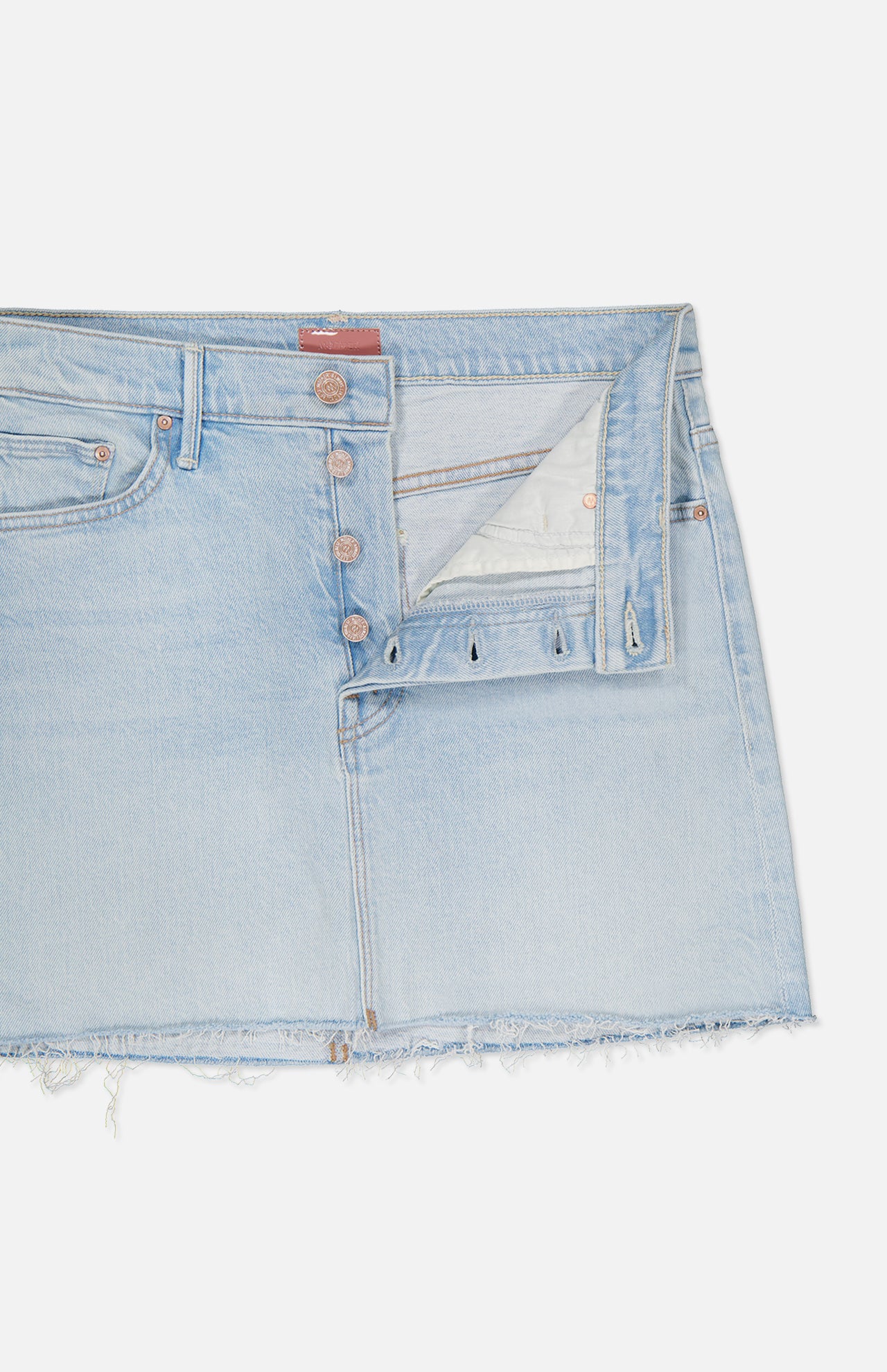 The Vagabond Mini Fray Skirt by MOTHER features a light blue, low-slung fit with a raw frayed hem, classic five-pocket styling, and a partially open button fly revealing white pocket lining. Shown against a white background.